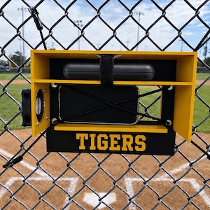 Peut inclure: Un tableau de bord de baseball jaune et noir avec le mot "TIGERS" en lettres dorées. Le tableau de bord est fixé à une clôture en treillis métallique surplombant un terrain de baseball. Un objet noir est fixé en haut du tableau de bord.