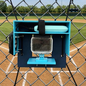 Peut inclure: Support de caméra bleu et noir fixé à une clôture en treillis métallique sur un terrain de baseball. Le support contient une caméra d'action transparente, un appareil rectangulaire bleu et une sangle noire. En arrière-plan, un terrain de baseball avec de l'herbe verte et un ciel bleu.