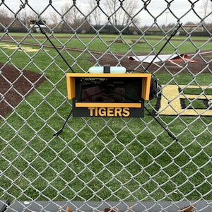 Könnte beinhalten: Ein gelb-schwarzes elektronisches Gerät mit dem Wort "TIGERS" ist an einem Maschendrahtzaun befestigt. Das Gerät hat einen Bildschirm und befindet sich vor einem Baseballfeld.