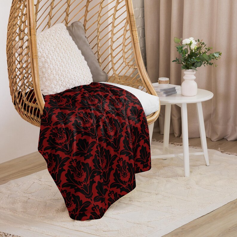 May include: A red and black patterned throw blanket draped over a wicker hanging chair. The blanket has a damask design with black floral patterns on a red background.