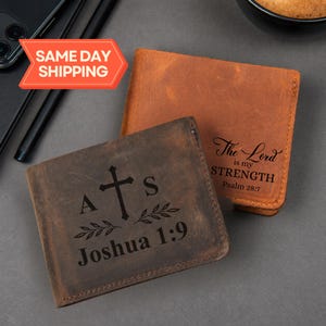 May include: Two leather wallets, one brown and one tan, are displayed on a gray surface. The brown wallet features a cross, initials, and the text "Joshua 1:9." The tan wallet has the text "The Lord is my Strength, Psalm 28:7."