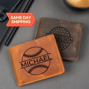 May include: Two leather wallets, one tan and one brown, are displayed. The tan wallet features a baseball design with the name "MICHAEL" engraved. The brown wallet has a golf ball design with the name "KELSON". The wallets are on a gray surface.