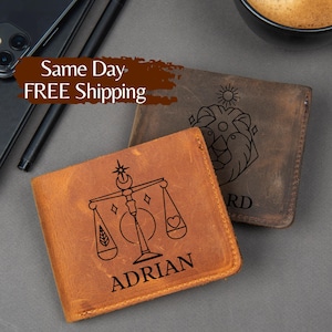 May include: Two leather wallets, one tan and one brown, are displayed. The tan wallet features a black Libra scale design with the name "ADRIAN" below. The brown wallet has a lion design. Both wallets are on a gray surface.