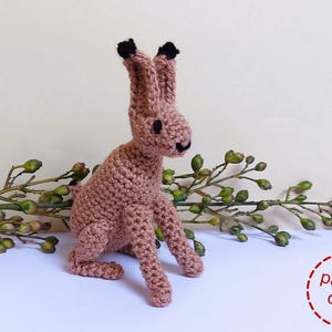 May include: Crochet pattern for a brown amigurumi hare with black ears and nose. The hare is sitting on a branch with green leaves.