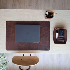 May include: A workspace setup seen from above, including a brown leather desk mat with a laptop, a pen holder with pencils, and a tray with a watch and wallet. The desk is light wood, and a wooden chair is in view.
