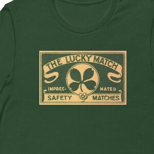 May include: A green t-shirt featuring a vintage-style graphic of a matchbox label. The label reads "The Lucky Match" with a four-leaf clover in the centre and the text "Impregnated Safety Matches".