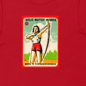 May include: A red t-shirt with a vintage matchbox label featuring a woman in a red and white outfit holding a bow and arrow. The label reads "Solo Match Works" and "Made in Czechoslovakia".