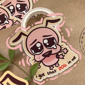 May include: A set of four pink and white cartoon dog stickers with a sad expression and a single tear. The stickers are on a brown paper background with a green leaf and the text "I got that DOG in me".