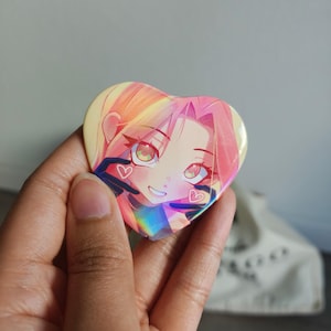 May include: Heart-shaped button featuring an anime-style illustration of a girl with pink hair and green eyes. The button has a holographic effect, with a rainbow sheen. Two small hearts are visible on the button.