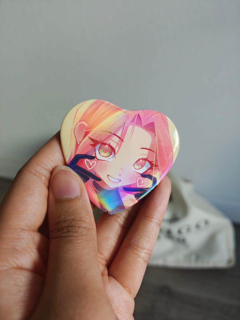 May include: Heart-shaped button featuring an anime-style illustration of a girl with pink hair and green eyes. The button has a holographic effect, with a rainbow sheen. Two small hearts are visible on the button.