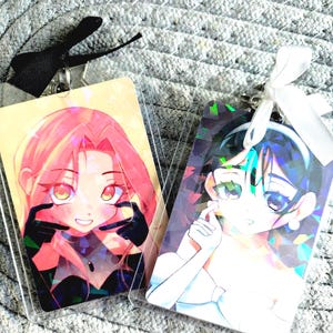 May include: Two anime-style character keychains. One features a pink-haired character with black gloves, the other a character with dark hair and a white outfit. Both are in clear plastic holders with black and white ribbons.