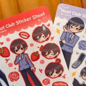 K&ouml;nnte beinhalten: Zwei Stickerb&ouml;gen mit Illustrationen im Anime-Stil. Ein rotes Blatt zeigt eine Figur in einem blauen Blazer, Erdbeeren, Sushi und Gesichter von Charakteren. Das andere ist lila und zeigt eine Figur im Anzug mit einem Telefon.