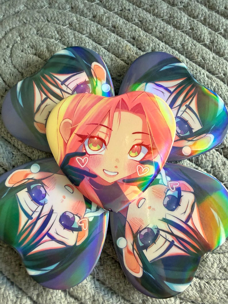 May include: Five heart-shaped anime character buttons. The central button features a character with pink hair and a smiling face. The surrounding buttons have a similar design with different hair colors and expressions. The buttons have a holographic sheen.