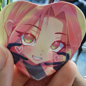 May include: Heart-shaped button featuring an anime-style illustration of a character with pink hair and green eyes. The character has a smiling expression and is surrounded by black gloves with white heart details.