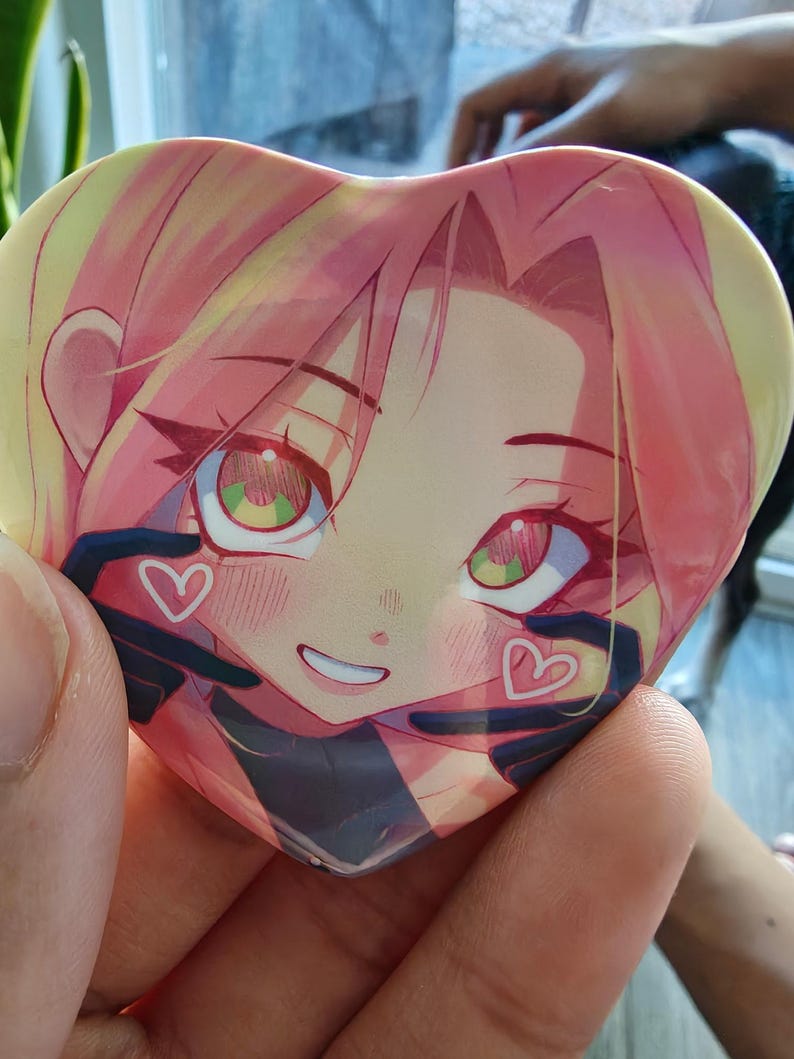 May include: Heart-shaped button featuring an anime-style illustration of a character with pink hair and green eyes. The character has a smiling expression and is surrounded by black gloves with white heart details.
