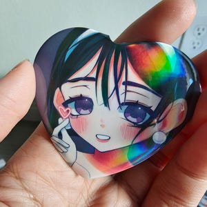 May include: Heart-shaped anime-style sticker featuring a girl with dark hair and purple eyes. The sticker has a holographic effect and a small pink heart. The girl is making a finger heart gesture.
