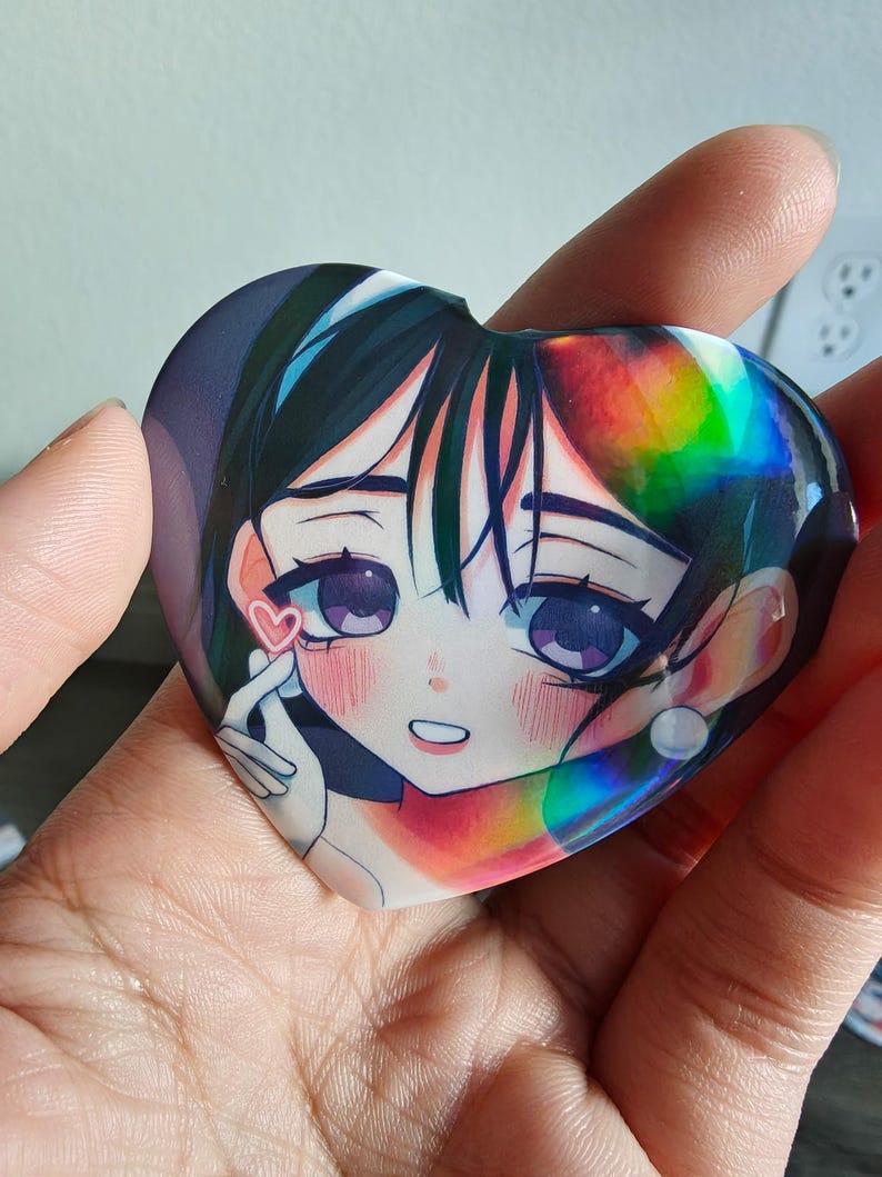 May include: Heart-shaped anime-style sticker featuring a girl with dark hair and purple eyes. The sticker has a holographic effect and a small pink heart. The girl is making a finger heart gesture.