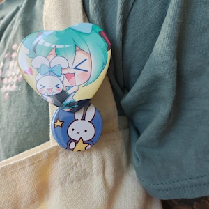 May include: Two colorful enamel pins are attached to a beige tote bag. The top pin is heart-shaped with a cartoon character with teal hair and a bunny. The bottom pin is round and blue with a white bunny and a yellow star.