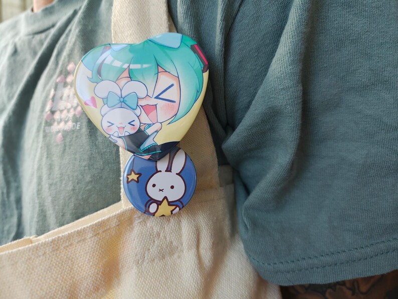 May include: Two colorful enamel pins are attached to a beige tote bag. The top pin is heart-shaped with a cartoon character with teal hair and a bunny. The bottom pin is round and blue with a white bunny and a yellow star.