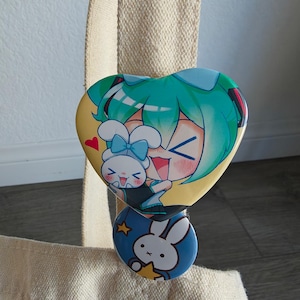 May include: A heart-shaped button featuring a cartoon character with teal hair and a bunny. Below, a round button with a bunny and stars. Both buttons are attached to a beige tote bag.