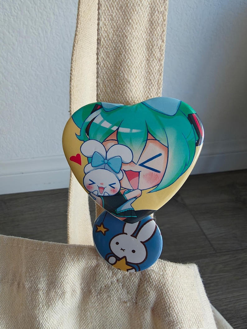 May include: A heart-shaped button featuring a cartoon character with teal hair and a bunny. Below, a round button with a bunny and stars. Both buttons are attached to a beige tote bag.