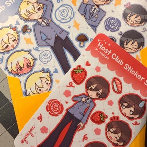 K&ouml;nnte beinhalten: Mehrere Stickerb&ouml;gen mit Illustrationen von Charakteren im Anime-Stil in Anz&uuml;gen. Die B&ouml;gen haben ein hellblaues und wei&szlig;es Farbschema mit dem Text "Host Club Sticker Sheet". Die Aufkleber enthalten Bilder von Erdbeeren, Rosen und anderen dekorativen Elementen.