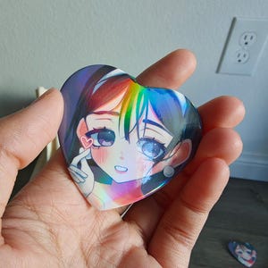 May include: A heart-shaped button featuring an anime-style illustration of a girl with rainbow-colored hair and large eyes. The button has a holographic finish, creating a shimmering effect. A smaller, similar button is visible in the background.