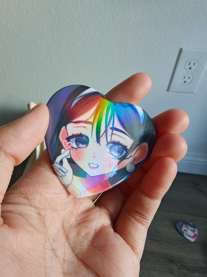 May include: A heart-shaped button featuring an anime-style illustration of a girl with rainbow-colored hair and large eyes. The button has a holographic finish, creating a shimmering effect. A smaller, similar button is visible in the background.