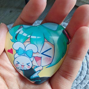 May include: Heart-shaped button featuring a cartoon illustration of two characters with teal hair and a white bunny. The button has a yellow background and a red heart. The button is held in a hand.