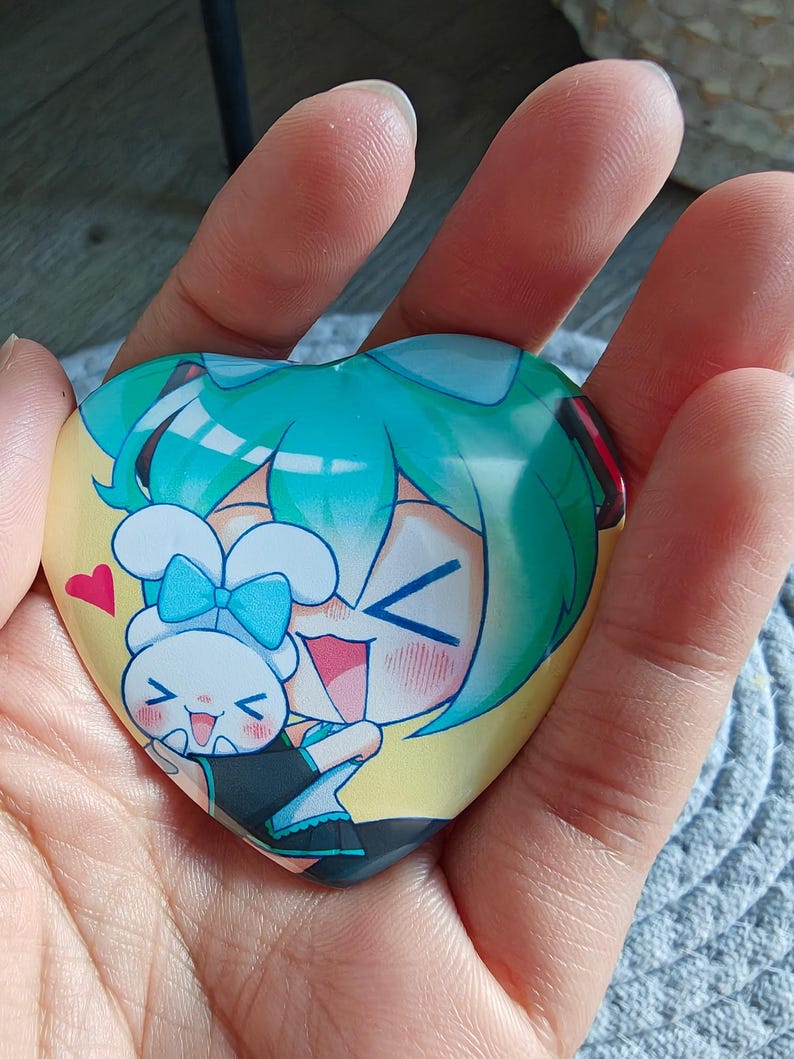 May include: Heart-shaped button featuring a cartoon illustration of two characters with teal hair and a white bunny. The button has a yellow background and a red heart. The button is held in a hand.