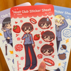 K&ouml;nnte beinhalten: Drei Stickerb&ouml;gen mit Illustrationen im Anime-Stil. Das zentrale Blatt ist rot und zeigt den Text "Host Club Sticker Sheet" und "01 - HARUHI". Die Aufkleber zeigen Charakterportr&auml;ts, Erdbeeren und Sushi.