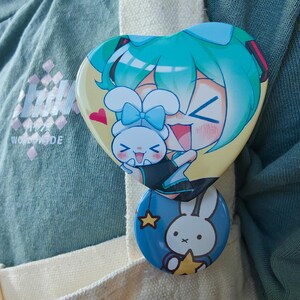 May include: Two colorful pinback buttons. The top button is heart-shaped with a cartoon character with teal hair and a white bunny. The bottom button is round with a white bunny and a yellow star on a blue background.