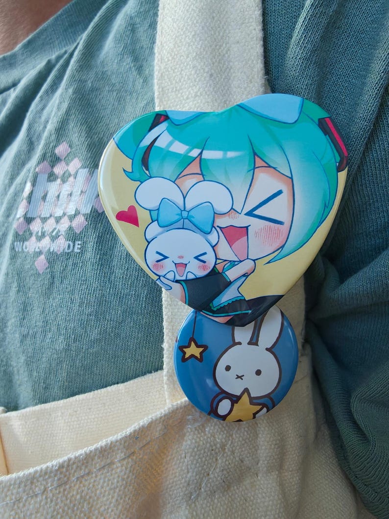 May include: Two colorful pinback buttons. The top button is heart-shaped with a cartoon character with teal hair and a white bunny. The bottom button is round with a white bunny and a yellow star on a blue background.