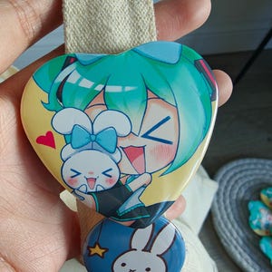May include: A heart-shaped button featuring a teal-haired anime character and a white bunny with a blue bow. Below, a round button with a white bunny and yellow stars on a blue background. The buttons are attached to a beige fabric.