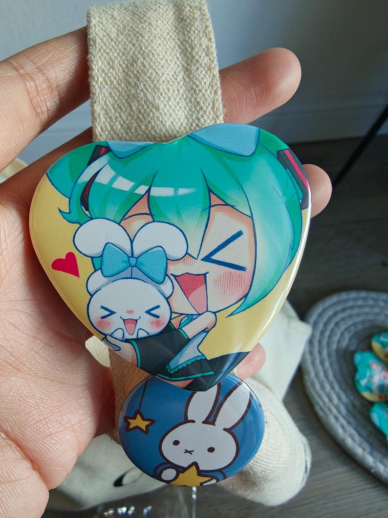 May include: A heart-shaped button featuring a teal-haired anime character and a white bunny with a blue bow. Below, a round button with a white bunny and yellow stars on a blue background. The buttons are attached to a beige fabric.