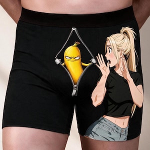 Peut inclure: Caleçon noir avec une impression graphique représentant une fermeture éclair ouverte révélant une banane jaune au visage grincheux. À côté de la fermeture se trouve l'illustration d'un personnage blond en queue de cheval portant un t-shirt noir.