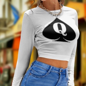 Könnte beinhalten: Ein weißes, langärmliges Crop-Top mit einem schwarzen Pik-Design, das ein großes "Q" in der Mitte zeigt. Das Top wird mit einer blauen Jeans getragen.