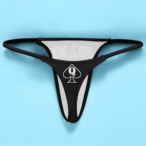 Op de afbeelding: Zwarte kanten string met de tekst "FILL ME" gedrukt op de achterkant en een wit schoppen-dame symbool op de voorkant.