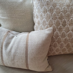 May include: Three decorative throw pillows with a beige and brown woven fabric. The pillows are arranged on a beige sofa.