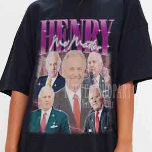 May include: Black t-shirt featuring a graphic of Henry McMaster with the text "HENRY McMaster" in a purple and white retro font. The graphic includes multiple images of the person. The shirt is a casual style.