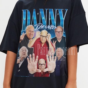 May include: Black t-shirt featuring a graphic of Danny DeVito with the text "DANNY DeVito" in a blue and white font. The graphic includes multiple images of Danny DeVito. The shirt is oversized.