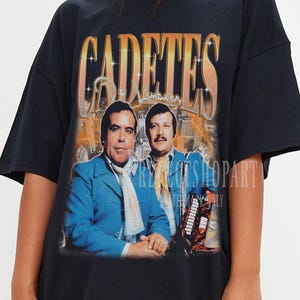 May include: Black oversized t-shirt featuring a vintage graphic of two men in blue suits with the word "CADETES" in gold lettering. The graphic has a faded, retro style, suggesting a music or entertainment theme.