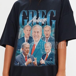 Puede incluir: Camiseta negra con un gráfico de un hombre con traje y el nombre "GREG" en letras grandes, azules y estilizadas. El gráfico incluye múltiples imágenes del hombre. La camiseta es de corte holgado.