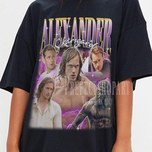 May include: Black oversized t-shirt with a graphic print of a collage of men's faces with the text "ALEXANDER Skarsgård" in purple and gold lettering.