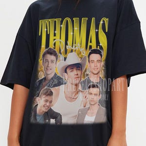 May include: Black oversized t-shirt featuring a vintage-style graphic with the name "THOMAS" in gold lettering. The graphic includes multiple images of a person wearing a cowboy hat and other individuals. The shirt is a casual, unisex style.