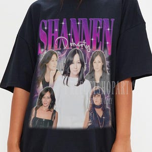 Könnte beinhalten: Schwarzes, übergroßes T-Shirt mit einer Grafik von Shannen Doherty und ihrem Namen in einem lila-weißen Design. Die Grafik enthält mehrere Bilder der Schauspielerin. Das Shirt wird von einer Person getragen.