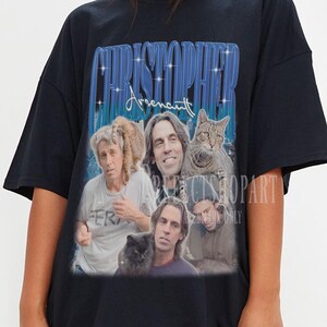 Könnte beinhalten: Schwarzes übergroßes T-Shirt mit einem Retro-Grafikdesign eines Mannes und einer Katze sowie dem Namen "CHRISTOPHER" in Blau. Das Shirt hat eine lockere Passform und kurze Ärmel. Das Design hat eine Vintage-Ästhetik.
