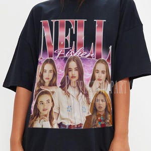 Puede incluir: Camiseta negra con el nombre "Nell Fisher" en degradado rosa y blanco. La camiseta tiene un gráfico de la imagen de una mujer debajo del texto. El estilo es holgado y de gran tamaño.