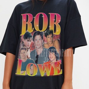 May include: Black oversized t-shirt with a graphic print of Rob Lowe. The graphic features a collage of images of Rob Lowe from different points in his career. The text "ROB LOWE" is printed in a gradient of pink, orange, and yellow.