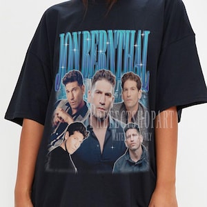 May include: Black oversized t-shirt featuring a vintage-style graphic of Jon Bernthal with the text "JON BERNTHAL" in a blue gradient font. The shirt has a crew neck and short sleeves. The graphic includes multiple images of the actor.